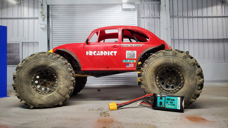 A remote control battery pack labeled 'Helios' placed on the ground next to a red RC car with a large tire, indicating the battery is for powering the vehicle.