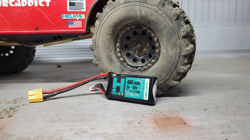 A remote control battery pack labeled 'Helios' placed on the ground next to a red RC car with a large tire, indicating the battery is for powering the vehicle.