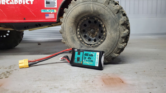 A remote control battery pack labeled 'Helios' placed on the ground next to a red RC car with a large tire, indicating the battery is for powering the vehicle.