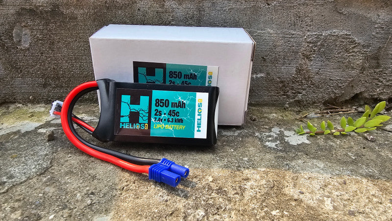 A 2S 850 MAH 45C LIPO battery with a blue label, connected to a red and black wire, placed on a stone surface.