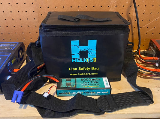 A black Helios RC Lipo Safety Bag with two pass-throughs for battery leads, a hand strap, and a removable shoulder strap, placed on a wooden surface with electronic components and wires around.