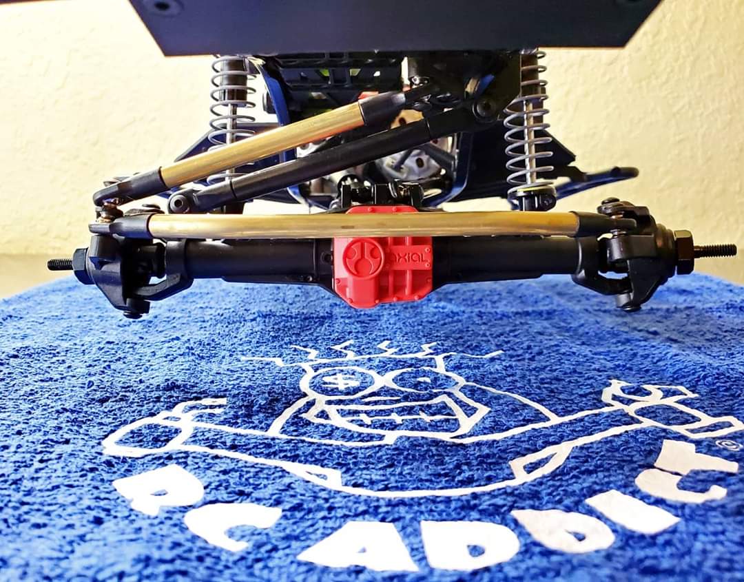 A brass steering link kit installed on a model vehicle, featuring high clearance links and a drag link, set against a blue backdrop with a white graphic.