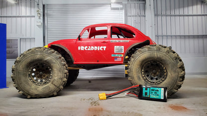 A red remote control car with 'H3Crazy' written on the side, featuring large off-road tires, parked inside a warehouse with a battery pack and charger on the floor.
